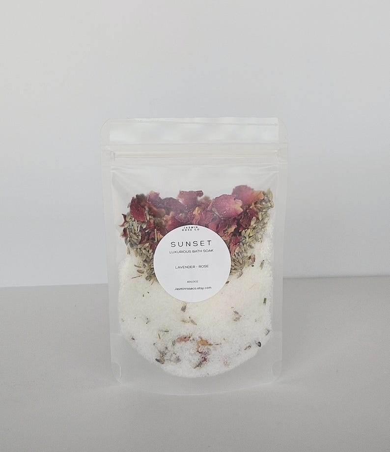 May include: A clear, resealable bag filled with a luxurious bath soak. The product is labeled "Sunset Luxurious Bath Soak Lavender - Rose." The soak contains dried rose petals, lavender buds, and white bath salts. The bag is displayed against a plain white background.