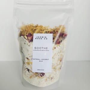 May include: A clear plastic bag filled with a white bath soak, dried flowers, and rose petals. The label reads "JASMIN ROSE CO" and "SOOTHE Luxurious Bath Soak, Oatmeal-Coconut Milk". The bag contains 10 oz of product.