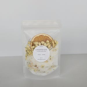 May include: A clear, resealable bag filled with white bath soak, dried orange slice, and jasmine buds. A white label reads "ENERGIZE LUXURIOUS BATH SOAK ORANGE - JASMINE". The bag is on a white background.