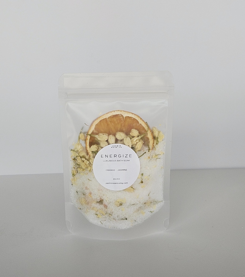 May include: A clear, resealable bag filled with white bath soak, dried orange slice, and jasmine buds. A white label reads "ENERGIZE LUXURIOUS BATH SOAK ORANGE - JASMINE". The bag is on a white background.
