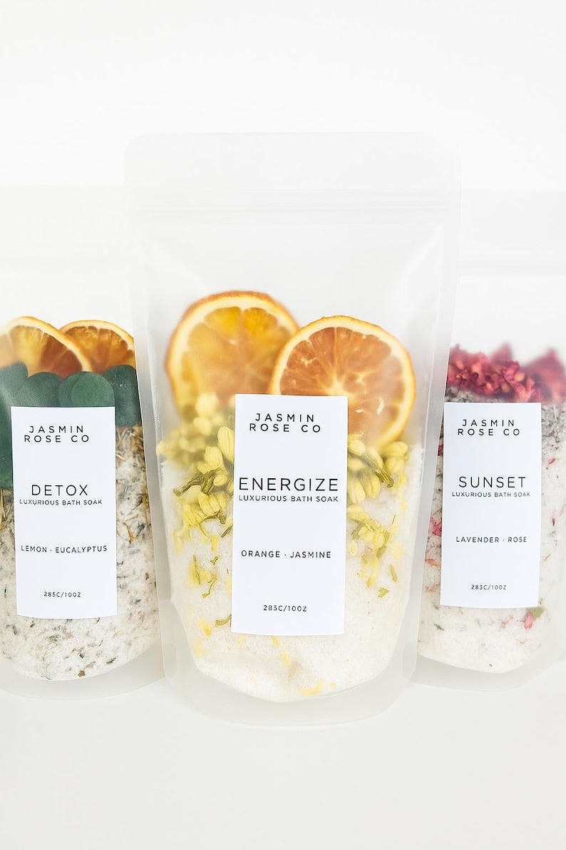 May include: Three clear pouches of bath soak with labels reading "Detox", "Energize", and "Sunset". Each pouch contains dried botanicals and citrus slices. The "Detox" pouch includes lemon and eucalyptus. The "Energize" pouch includes orange and jasmine. The "Sunset" pouch includes lavender and rose.