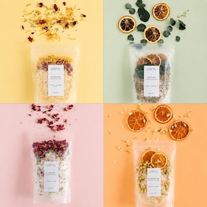 May include: Four clear pouches of bath products with labels, each on a different colored background. The pouches are labeled "Soothe", "Detox", "Sunset", and "Energize". Dried flowers and orange slices are arranged around the pouches.