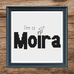 May include: A white cross-stitch embroidery with black text that reads "I'm a Moira" and a small white bird with a gold halo above the word Moira.