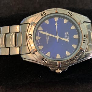 May include: A silver-tone analog wristwatch with a deep blue circular dial. The watch features a metallic link bracelet, a rotating bezel with engraved numbers, and the text Alpha Centauri, Quartz, and Water Resistant printed on the face.