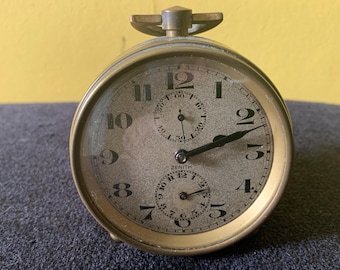Könnte beinhalten: Eine antike Zenith Taschenuhr mit goldfarbenem Gehäuse und grauem Zifferblatt. Die Uhr hat schwarze Zeiger und Ziffern mit zwei kleineren Zifferblättern. Die Uhr ist vor einem gelben Hintergrund platziert.