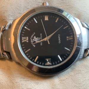 May include: A silver men's wristwatch with a black face and silver hands. The watch face has Roman numeral hour markers and the text "Beverly Hills Polo Club Quartz".