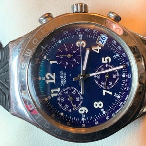 May include: A silver and black chronograph wristwatch with a blue face and white markings. The watch has a date window at the 3 o'clock position and the words 'swatch swiss' printed on the face.