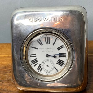 May include: A silver clock with a white face and black Roman numeral hour markers. The clock is in a silver case with the inscription "Quo Vadis" on the top.
