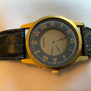 Può includere: Un orologio dorato con un cinturino in pelle nera e un quadrante rotondo con numeri romani. Il quadrante dell'orologio ha un piccolo quadrante circolare grigio con la parola "QUARTZ" stampata su di esso.