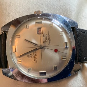 May include: A vintage Mortima Super De Luxe wristwatch with a silver-colored face, black leather band, and a date display. The watch has a silver-colored bezel and a crown. The watch face displays the text "Mortima 17 Jewels Super Automatic".