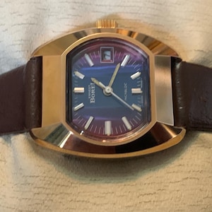Puede incluir: Un reloj vintage dorado con esfera rectangular y esfera azul oscuro. El reloj tiene una correa de cuero marrón. La esfera muestra la fecha y la marca "Borel".