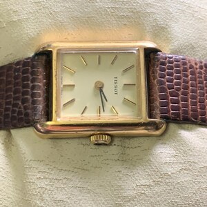 May include: A gold Tissot watch with a square face and a light-coloured dial. The watch has gold hands and markers, and a brown, textured leather strap. The watch is a classic style.