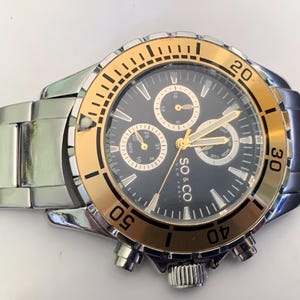 Puede incluir: Un reloj cronógrafo SO & CO New York plateado y dorado. El reloj presenta una esfera negra con agujas y marcadores dorados, tres subesferas y una ventana de fecha. El bisel dorado tiene marcas negras y el reloj tiene una correa de acero inoxidable plateado.