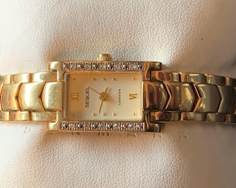 Può includere: Un orologio color oro con un quadrante rettangolare e una lunetta tempestata di diamanti. Il quadrante dell'orologio è bianco con indici orari in numeri romani neri e le parole "SEQUEL LONDON" stampate su di esso.