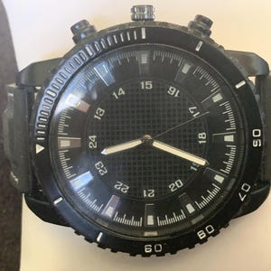 Puede incluir: Reloj de pulsera negro con esfera redonda y correa de cuero negro. La esfera del reloj tiene números y manecillas blancas, con un centro texturizado. El reloj tiene tres botones y un bisel con marcas.