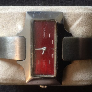 Puede incluir: Un reloj de pulsera plateado con una esfera roja y agujas blancas. La esfera del reloj tiene la palabra "SICURA" y "17 jewels incabloc" impresas.