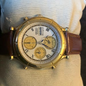 May include: A gold Seiko Quartz Chronograph watch with a white face, Roman numerals, and three sub-dials. The watch has a brown leather strap and the day of the week is displayed. The watch is on a cream-coloured surface.