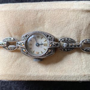 May include: A silver wristwatch with a white face and black Roman numeral markings. The watch has a silver band with marcasite stones.
