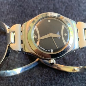 Könnte beinhalten: Eine Swatch-Uhr mit schwarzem Zifferblatt und silberfarbenem Metallarmband. Die Uhr hat ein rundes Zifferblatt mit silbernen Zeigern und Stundenmarkierungen. Das Armband ist im Manschettenstil. Die Uhr ist mit "Swatch AG 2001" gekennzeichnet.