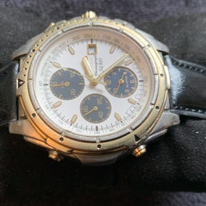May include: A silver and gold chronograph wristwatch with a black leather band. The watch face is white with black markings and has three sub-dials. The watch has a tachymeter scale around the outer edge of the face.