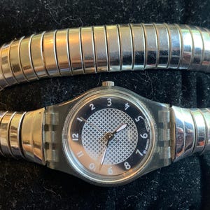 Könnte beinhalten: Eine Swatch-Armbanduhr mit rundem, transparentem Kunststoffgehäuse und schwarzem Zifferblatt mit weißen arabischen Ziffern. Die Mitte des Zifferblatts hat ein weißes Kreuzschraffurmuster. Die Uhr hat ein flexibles, silberfarbenes Metallarmband.