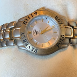 May include: A silver and gold-toned Beverly Hills Polo Club watch. The watch has a silver face with gold hands and a gold bezel. The watch band is silver and gold-toned. The words "Beverly Hills Polo Club" are inscribed around the watch face.