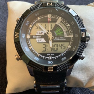 Puede incluir: Reloj de pulsera Shark negro y plateado con pantalla digital y bisel de brújula. La esfera del reloj muestra la hora 3:10:45 y las palabras "Dual Time Alarm Chrono".