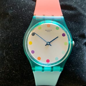 Puede incluir: Un reloj Swatch turquesa y blanco con una esfera blanca y puntos de colores. El reloj tiene una correa rosa.