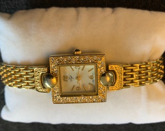 Può includere: Un orologio color oro con quadrante quadrato e cinturino in metallo dorato. Il quadrante è circondato da piccoli cristalli trasparenti. Il quadrante dell'orologio ha uno sfondo bianco con indici orari in numeri romani neri e lancette nere.