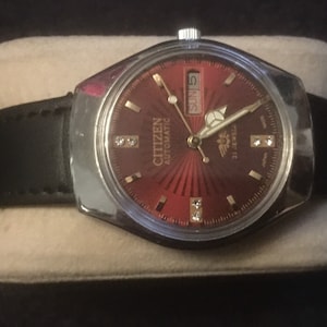 Op de afbeelding: Een Citizen automatisch polshorloge met een rode wijzerplaat en een zwart leren bandje. De wijzerplaat van het horloge heeft gouden accenten en een datumscherm dat "SUN 5" aangeeft.