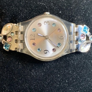 Könnte beinhalten: Eine Armbanduhr aus klarem Kunststoff mit einem silberfarbenen Zifferblatt, das die Zahlen 3, 6, 9 und 12 zeigt. Das Zifferblatt ist mit kleinen, bunten runden Kristallen verziert. Das Armband besteht aus silberfarbenen Gliedern mit blauen, rosa und klaren Kristallen.