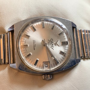 May include: A silver wristwatch with a white face and black hands. The watch has a silver band and says "Le Chlore" and "Camy Geneva 17 Jewels" on the face.