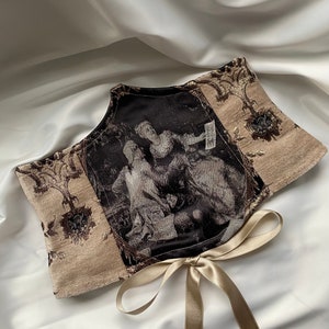 Op de afbeelding: Een decoratief halssieraad met een klassiek schilderij in zwart-wit, omlijst door beige stof met een bloemmotief. Het is vastgebonden met een bijpassend beige satijnen lint. De achtergrond is een witte, zijdeachtige stof.