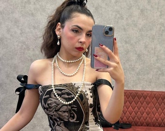 May include: A woman wearing a black and white corset with a Medusa print. She is wearing a pearl necklace and earrings. She has her hair pulled back in a ponytail with a black ribbon.