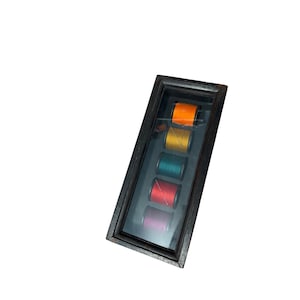 Può includere: Una teca rettangolare in legno scuro con frontale in vetro contenente cinque rocchetti di filo da cucito disposti verticalmente. I colori dei fili dall'alto verso il basso sono arancione brillante, giallo oro, verde ottanio, rosso e magenta.
