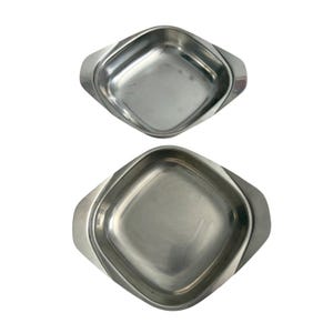 May include: Two square stainless steel serving dishes with rounded corners and integrated side handles. The dishes have a polished silver finish and a smooth, reflective surface. One dish is noticeably larger than the other, both featuring a simple, open design for food presentation.