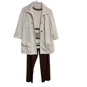 May include: A cream-colored jacket with toggle closures and two front pockets, worn over a white sweater featuring a brown geometric pattern at the neckline and midsection, paired with solid dark brown trousers on a white hanger.