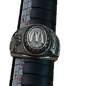 May include: A silver-toned metal ring featuring a central oval emblem with a raised letter M logo. The ring is positioned on a black plastic ring sizer tool with white markings, showing the band size between 7 and 8 on the scale.