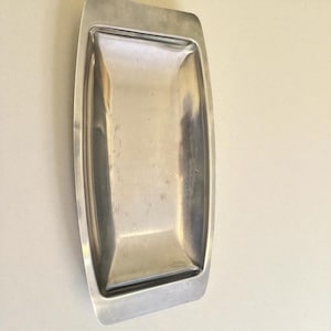May include: A rectangular, stainless steel serving tray with a slightly curved, raised edge. The tray has a polished, reflective surface and a simple, minimalist design. The tray is empty and appears to be vintage.