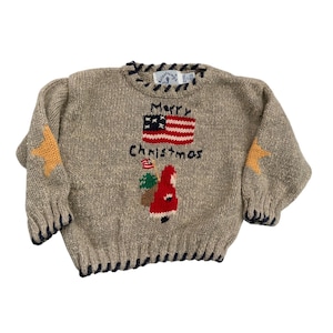 May include: Tan knit Christmas sweater with a flag design that says "Merry Christmas". The sweater has a red, white, and blue flag, a Santa Claus design, and yellow stars on the sleeves.