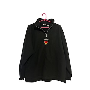 May include: Black pullover sweatshirt with a quarter-zip neckline. The sweatshirt features a small embroidered emblem with the German flag colors: black, red, and gold. The emblem is located on the left chest area. The sweatshirt is hanging on a pink hanger.