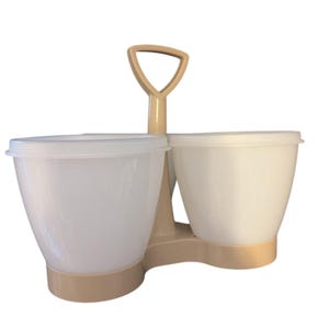 May include: A beige and white plastic mixing bowl set. The set includes two bowls with lids, connected by a beige handle. The bowls are white and have a rounded shape. The handle is shaped like a shovel.