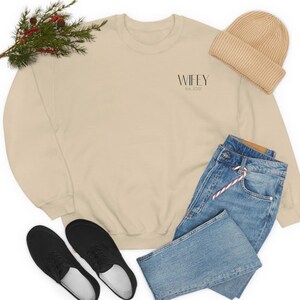 Puede incluir: Una sudadera beige con el texto "WIFEY Est. 2022" en la parte delantera. La sudadera se combina con unos vaqueros azules, una boina beige y unas zapatillas negras.