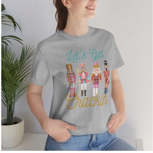 May include: A grey t-shirt featuring a graphic of four nutcracker figurines and the text "Let's Get Crackin'" in teal and gold lettering.