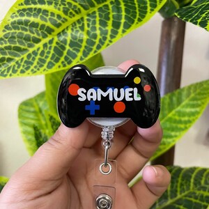 May include: Black and white game controller badge holder with the name "SAMUEL" in white letters. The badge holder has a clear plastic retractable cord and clip.