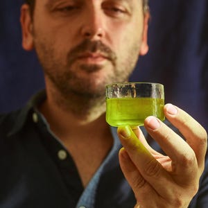 以下が含まれることがあります： 人が、明るい緑色の液体で満たされた小さなガラスのショットグラスを持っています。液体は透明で、わずかに黄色がかった色合いをしています。