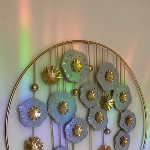 May include: A gold metal wall decor with a circular frame and a design of white flowers with gold centers. The flowers are arranged in a circular pattern and are connected by gold metal rods.