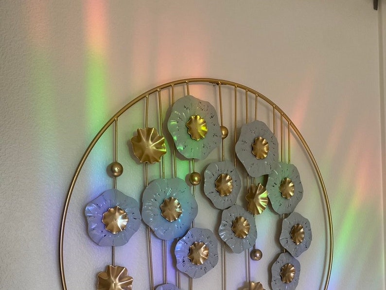 May include: A gold metal wall decor with a circular frame and a design of white flowers with gold centers. The flowers are arranged in a circular pattern and are connected by gold metal rods.