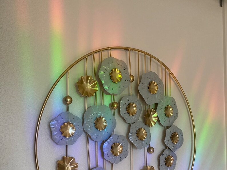 May include: A gold metal wall decor with a circular frame and a design of white and gold flowers. The flowers are arranged in a cascading pattern and have a textured surface.