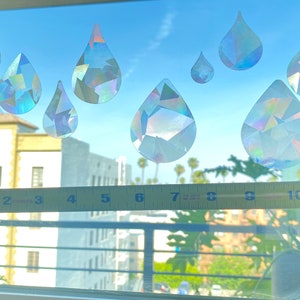 May include: A window with seven iridescent rainbow-colored raindrop stickers on it. A yellow measuring tape is placed on the window sill.